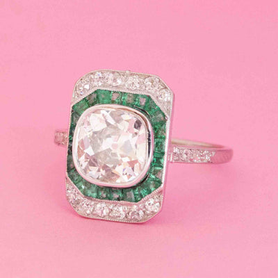 Art Deco dinner ring with 2.11ct old mine cut diamond, emerald halo, and diamond accents in platinum on pink background