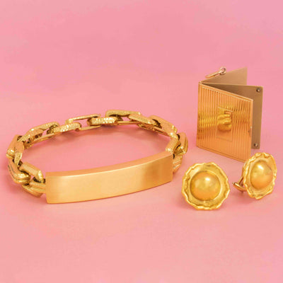Designer gold jewelry set with textured bracelet, vintage clip-on earrings, and collectible compact case on pink background