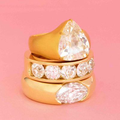 Stack of JbyG gold rings featuring antique-cut diamonds in bold bezel and half-hoop settings, shown against a pink background