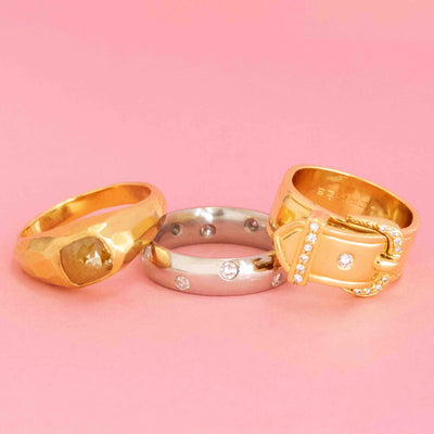 Trio of modern rings in gold and platinum featuring unique shapes, diamonds, and a Hermès buckle design, shown on a pink background.