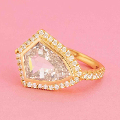 East-west shield cut diamond halo ring in 18kt yellow gold with peachy center stone on pink background