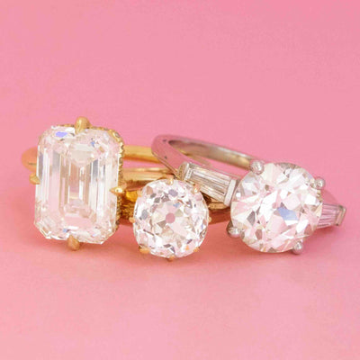 Trio of vintage and antique engagement rings featuring emerald cut, old mine cut, and old European cut diamonds on a pink background.