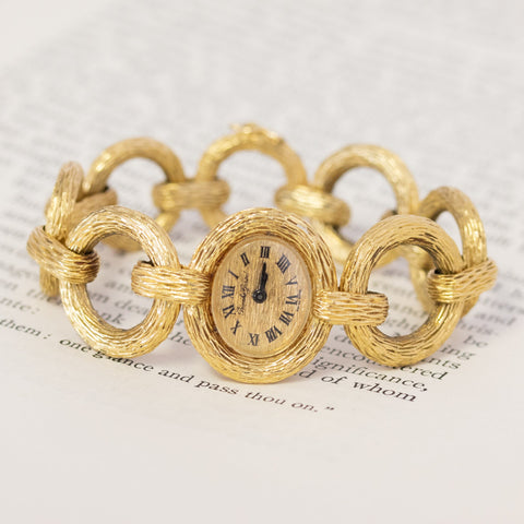 A vintage Bueche Girod gold chain link watch with intricate engravings on the links, displayed on an open book.