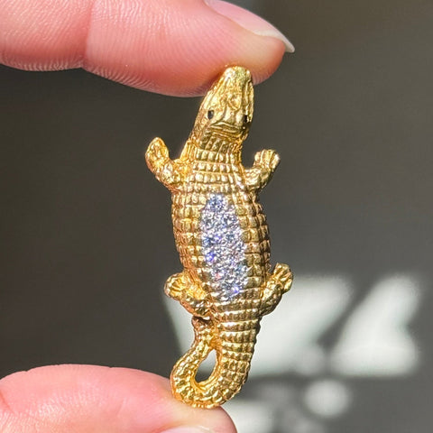 A 18kt gold crocodile or alligator-shaped pendant with a textured scale finish and a clear rhinestone-embellished belly, held between two fingers against a dark gray background.