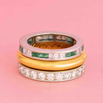 Stack of vintage and antique bands including an emerald and diamond eternity ring, classic gold band, and engraved diamond band on a pink background