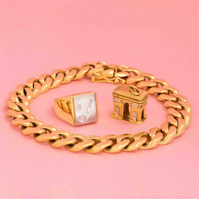 Vintage men’s gold jewelry including a chunky chain bracelet, a golf-themed signet ring, and an Arc de Triomphe gold charm on pink background