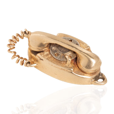 A vintage-style rotary telephone pendant or charm in a warm gold, featuring a coiled cord and dial, photographed on a clean white background with a faint reflection below.