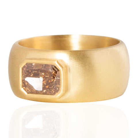 A wide, chunky yellow gold ring featuring a large, faceted rectangular brown radiant cut diamond in a bezel setting, shown on a clean white background with a soft reflection below.
