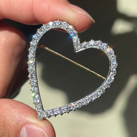 A heart-shaped platinum brooch paved with small clear stones, held between fingers against a softly blurred light background.