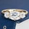 A yellow gold three-stone ring with a large oval center diamond and two round side diamonds, displayed on a textured navy blue surface.