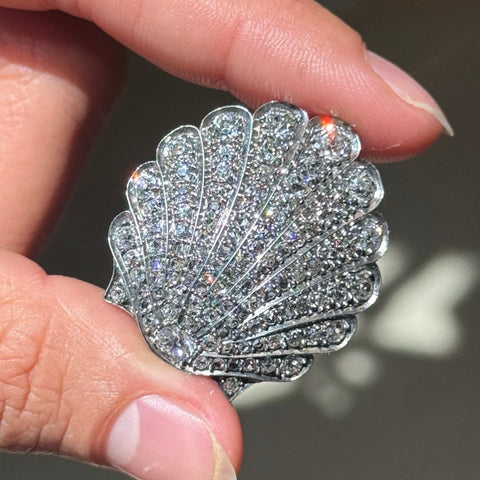 A close-up of a hand holding a scallop-shaped silver brooch or pin covered in sparkling clear diamonds.