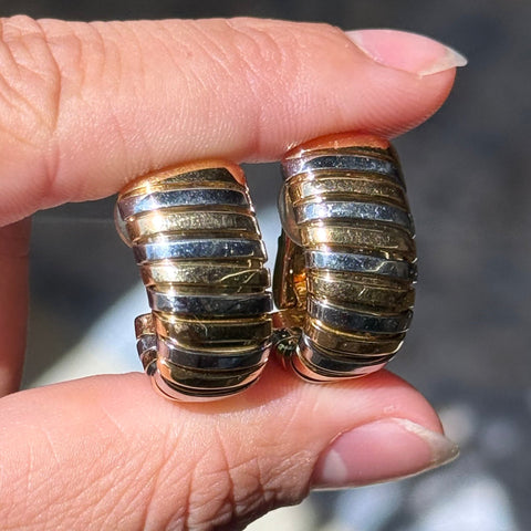 A close-up of a hand holding a pair of chunky hoop earrings with alternating horizontal bands of gold and silver-toned metal.