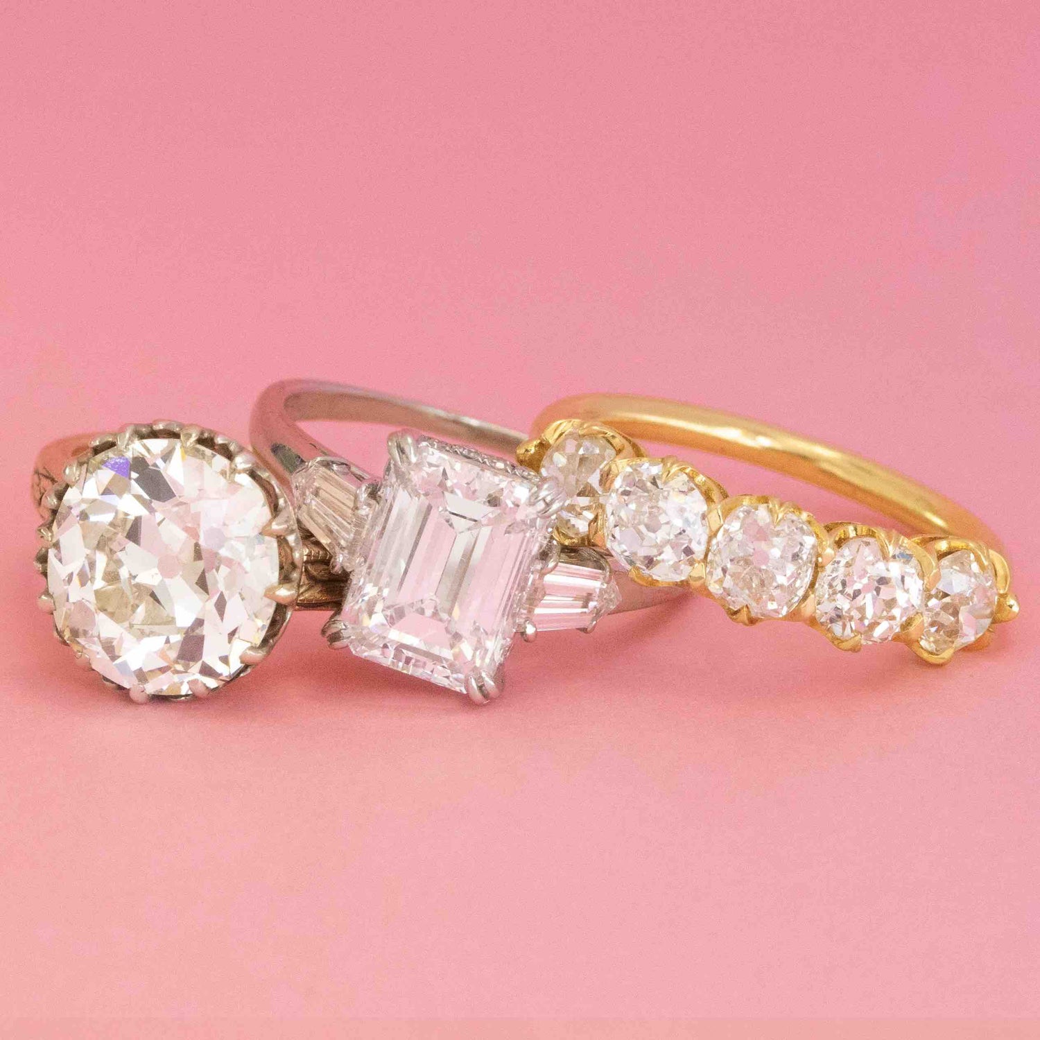 Trio of vintage and antique diamond rings in platinum and gold, featuring emerald cut, old European cut, and five-stone designs against a pink background.