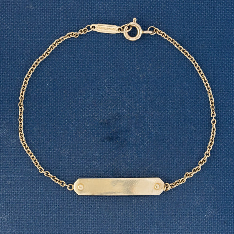 A delicate designer Tiffany & Co. gold chain bracelet with a polished rectangular bar pendant, displayed on a textured blue background.