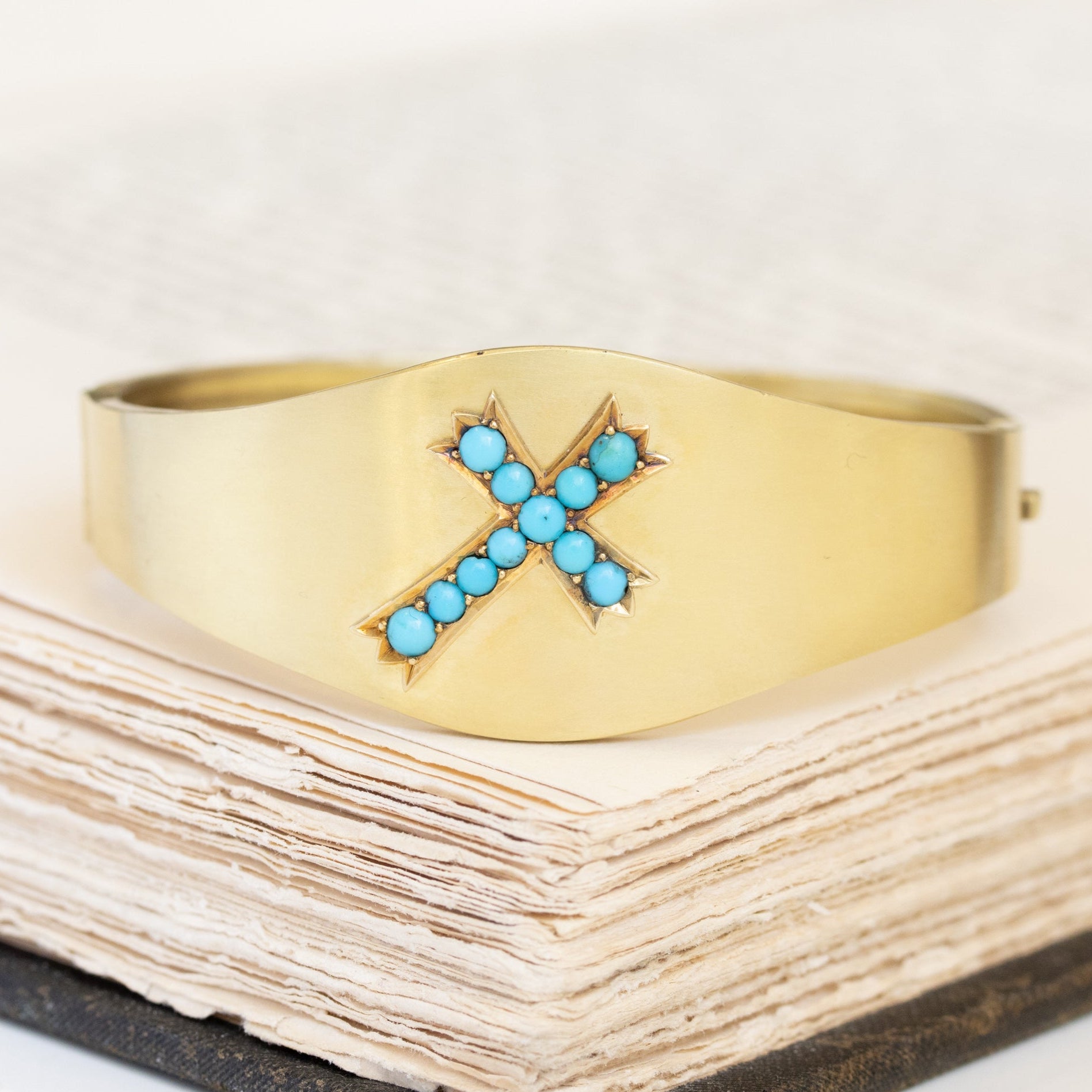 An antique turquoise and yellow gold bangle bracelet with a cross motif design, displayed on a book.